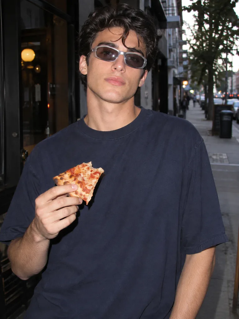 Un jeune homme portant des lunettes de soleil Y2K et un t-shirt bleu marine tout en tenant une part de pizza