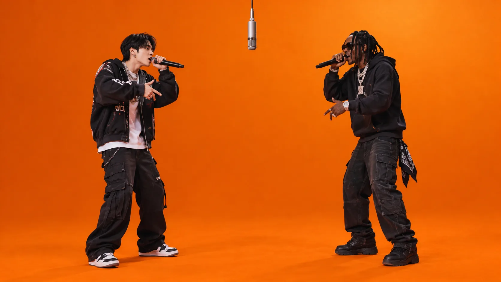 Two rappers in black streetwear performing with mics under a hanging mic in an orange booth